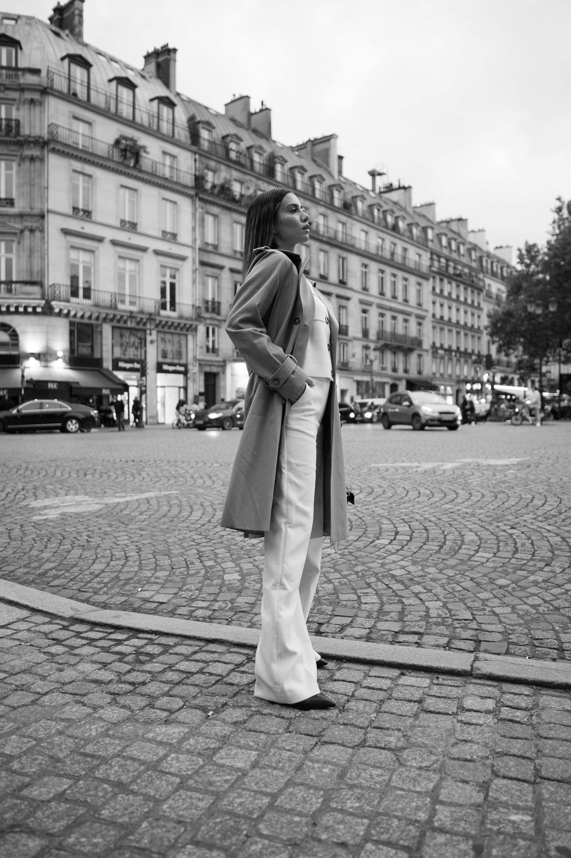 Une femme en trench et pantalon blanc dans une rue pavée de Paris, devant des immeubles haussmanniens